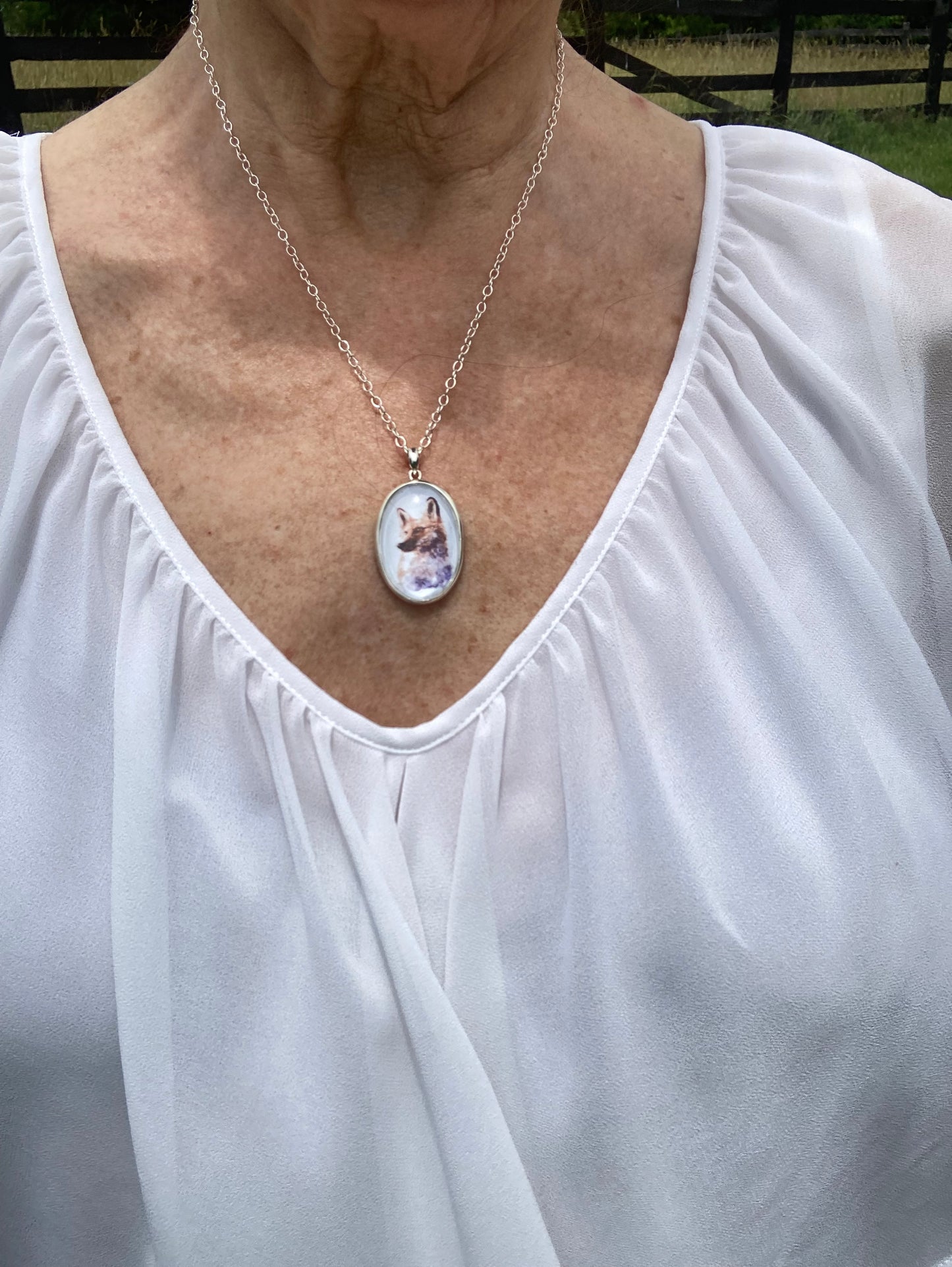 Sterling silver fox pendant featuring a reproduction of an original watercolor by Salina Ramsay beneath a crystal-clear dome. The artwork captures the alert expression and natural beauty of a fox in soft woodland tones, creating a collectible wearable work of art inspired by nature and country life. Shown worn for scale. 